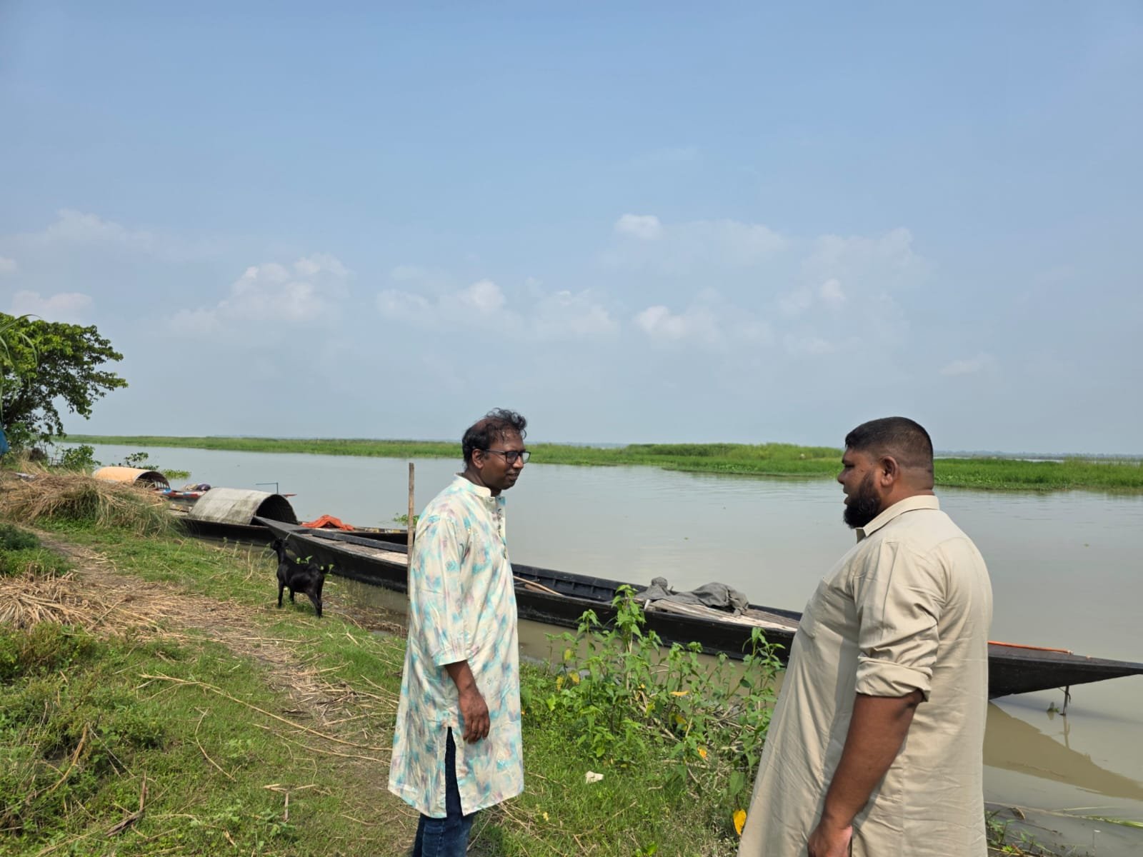 Two men talking beside the river, fishing boats moored along the bank behind them